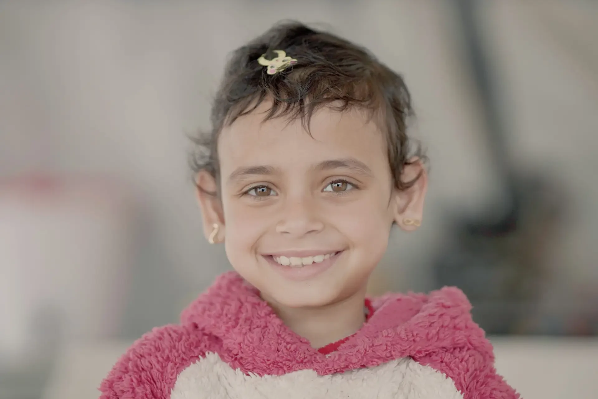 A young girl with short, dark brown hair smiles directly at the camera. She wears a pink and cream-colored fleece hoodie, small gold earrings, and a decorative hair clip. The background is softly blurred, keeping the focus on her face.